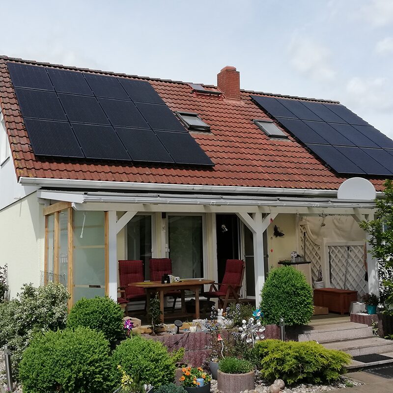 Weißes Wohnhaus mit zwei Dachflächen voller Solarpaneele, davor bunte Pflanzen und eine überdachte Terrasse.