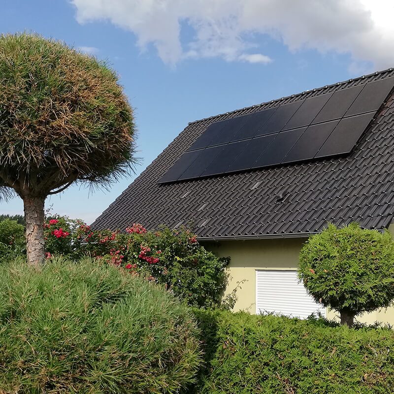 Gelbes Einfamilienhaus mit dunklem Ziegeldach, auf dem eine Photovoltaikanlage installiert ist, davor ein runder Baum.