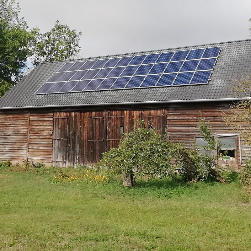 Verwitterte Scheune mit Solarpaneelen auf dem Dach, umgeben von Wiese und Bäumen.